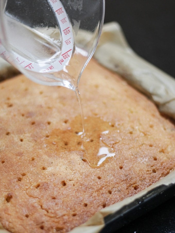 Cooked Gin and Tonic Cake bar in the baking tin lined with baking paper, drizzling on Gin and Tonic syrup from a plastic measuring jug.