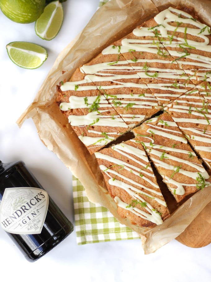 Overhead photo of gin and tonic cake topped with white chocolate and lime with a bottle of Hendricks gin laying next to the cake. With lime wedges and a grey check tea towel.