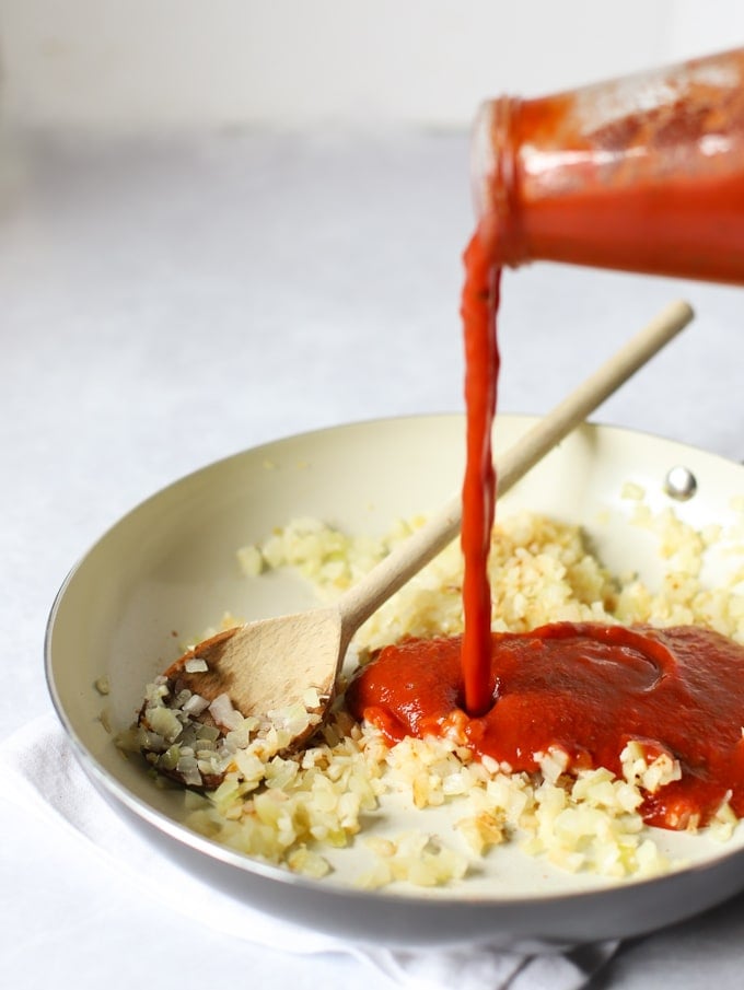 Fried onions in a frying pan with passata being poured in for step 1 in the recipe for Gnocchi Bake.