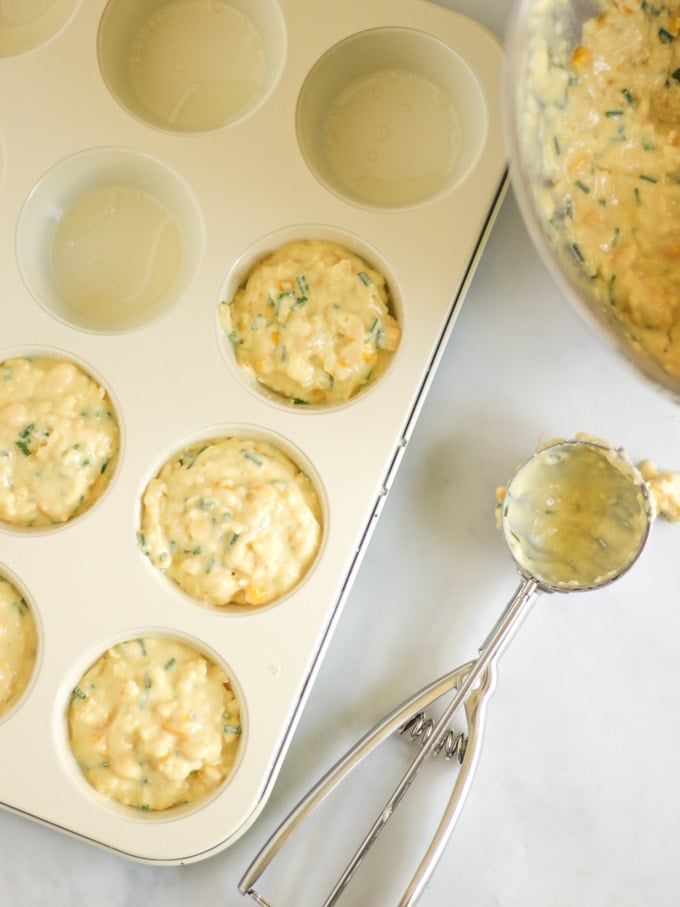 A bowl of savoury muffin batter mixture and an ice cream scoop, with a muffin tray half filled with the batter.