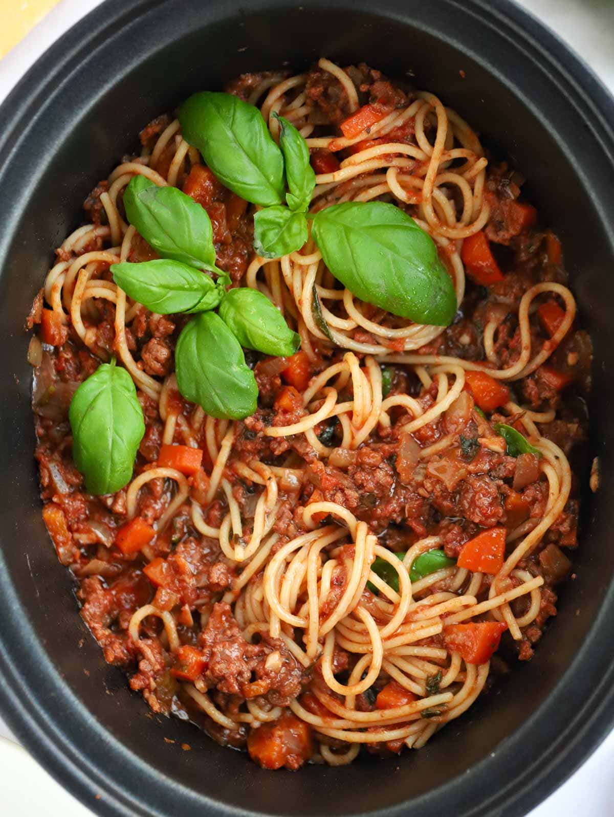 Slow cooker bolognese sauce in a slow cooker pan, mixed with cooked spaghetti and topped with basil.