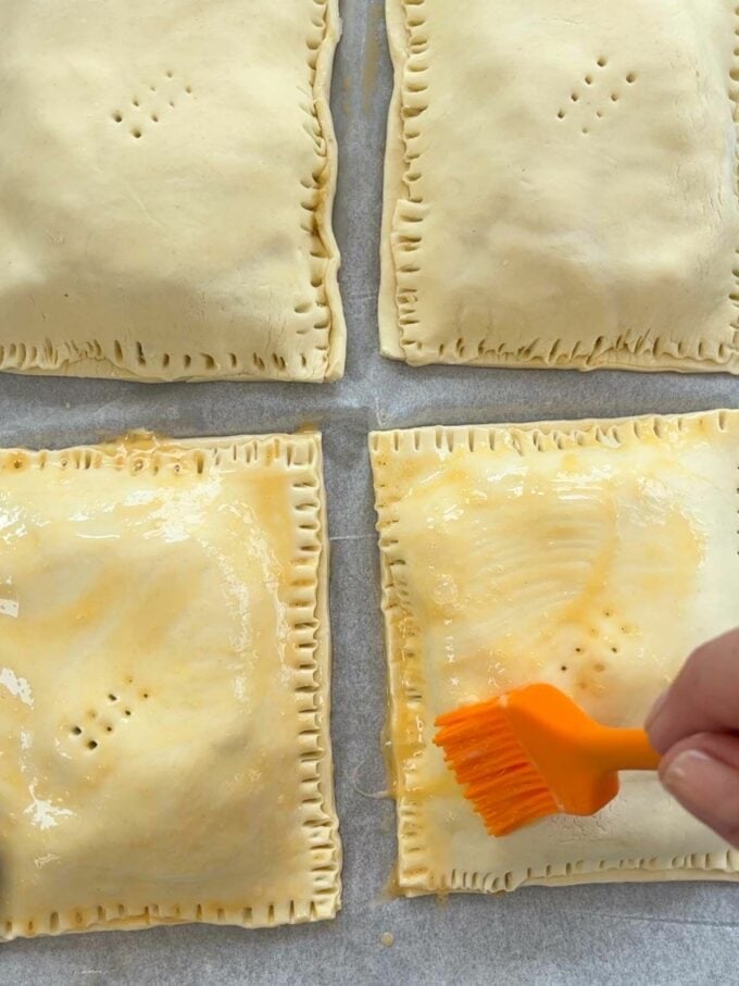 Four uncooked pasties, with a hand brushing egg wash on top of them.