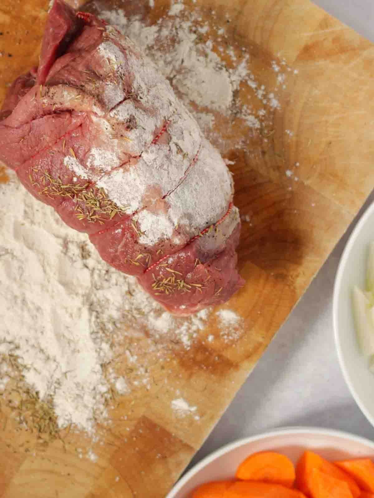 Step 1 in the recipe for how to make a beef joint in the slow cooker. A raw joint being rolled in flour on a wooden board.
