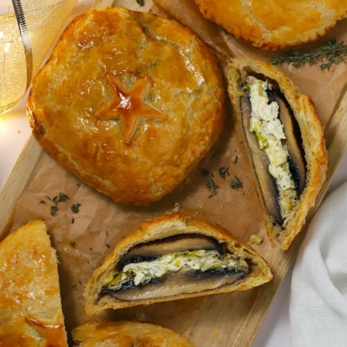 Four puff pastry mushroom wellingtons on a long copping board, with one sliced and on its side to show the filling inside.