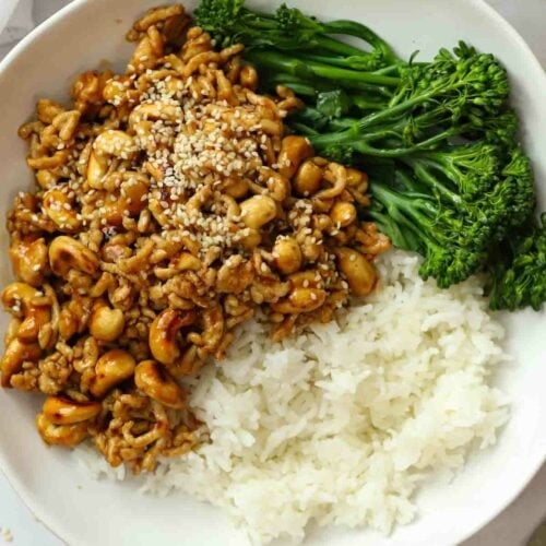 A plate on a white table filled with cooked cashew chicken stir fry, with a side of white rice and broccoli.