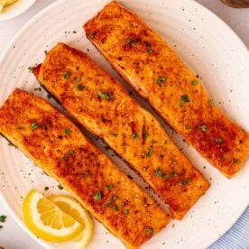 A white plate with three salmon fillets on and half moons of lemons to the side.