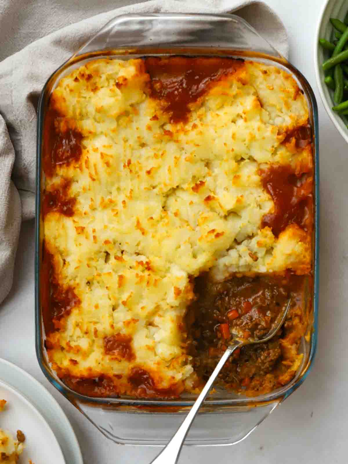 A cooked cottage pie in an oven dish, being served with a spoon, with one portion missing.