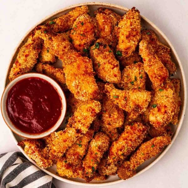 A round plate filled with golden, crispy Chicken Goujons, also named Chicken Tenders, with a bowl of ketchup for dipping.