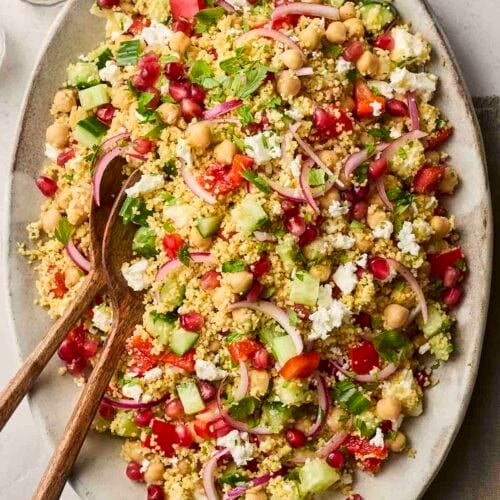 A colourful plate of salad with couscous, chickpeas and lots of other ingredients, with a pepper mill to the side and salad utensils to serve.