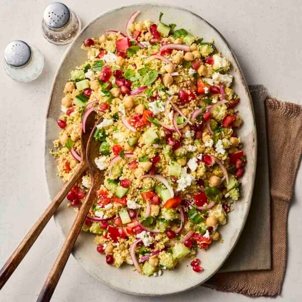 A platter with a colourful couscous salad on, sitting on a white counter with salad servers ready to dish out.