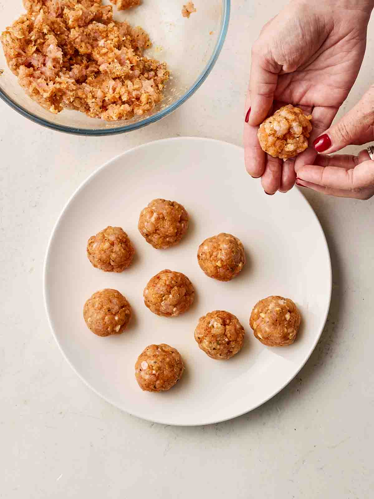 A bowl of chicken mince with a white plate to the side with meatballs on, and two hands rolling one up.