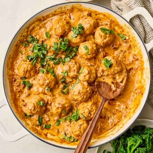 A large pan filled with creamy chicken meatballs for Marry Me Meatballs recipe, on table with a serving spoon and a bowl of mash and broccoli to serve.