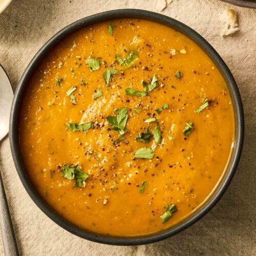 A bowl of orange coloured soup on a table with chopped coriander over the top.