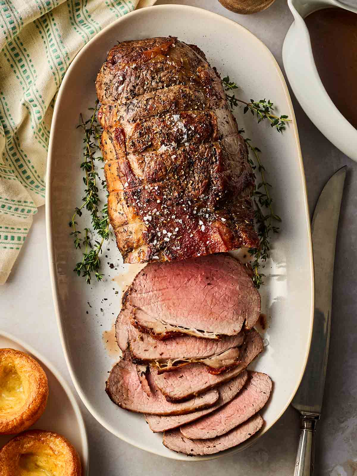 A perfectly cooked roast beef joint on a white platter, with sprigs of thyme around the sides, on a table ready to be served.