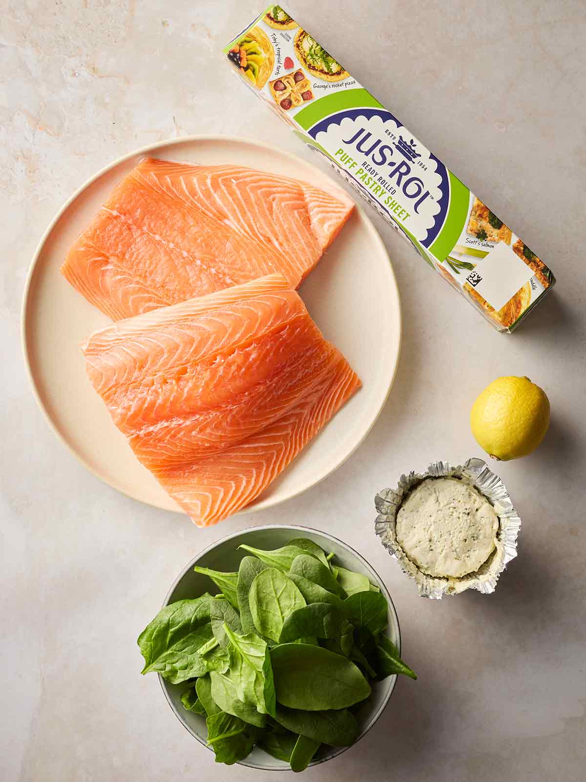Raw ingredients laid out on a white counter, including salmon, spinach, lemon, puff pastry and boursin.