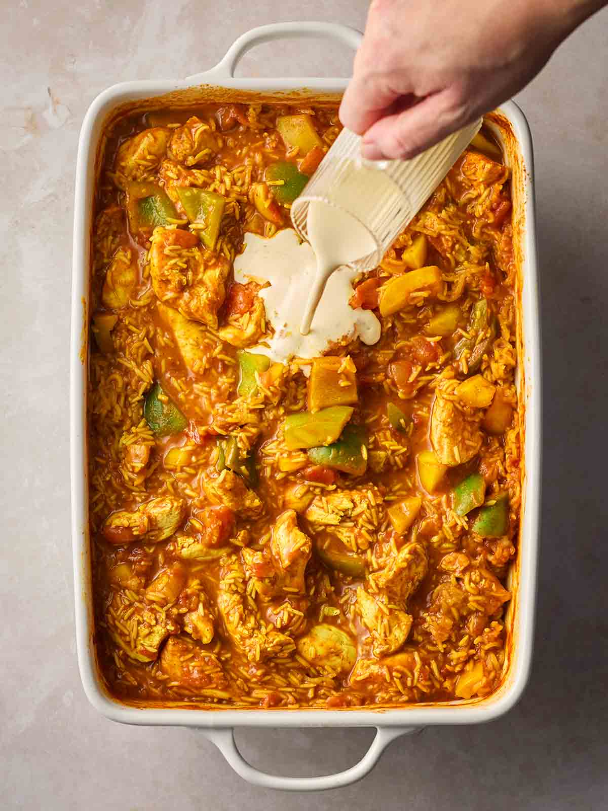 A hand pouring a jug of cream into a dish of cooked chicken curry and rice.