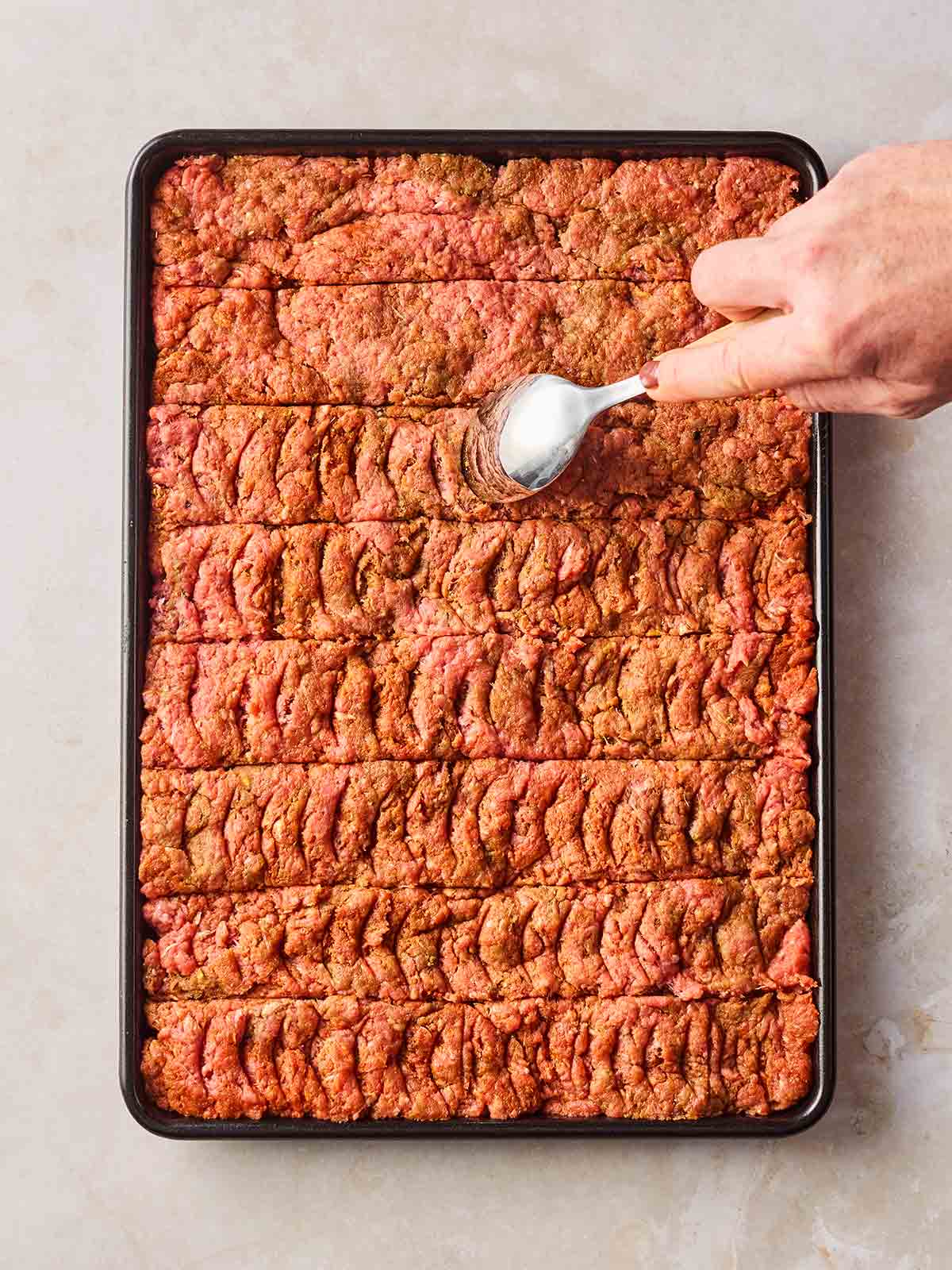 An oven sheet with a mince mixture pressed into it, and a hand scoring the meat with a spoon.