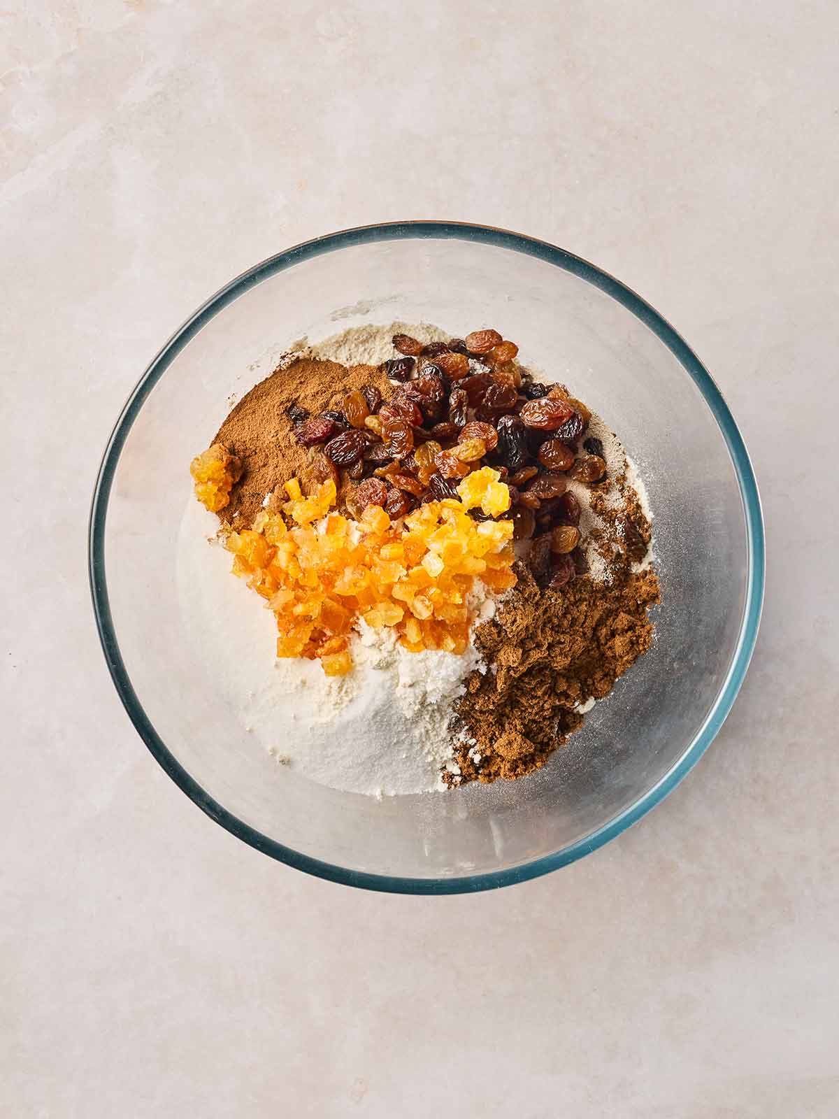 A glass bowl with spices, flour, sugar and dried fruit in, ready to mix.