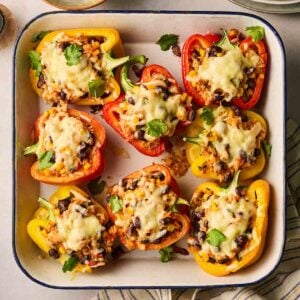 A close up of halved peppers in a roasting dish, stuffed with rice and veggies, with melted cheese on top.