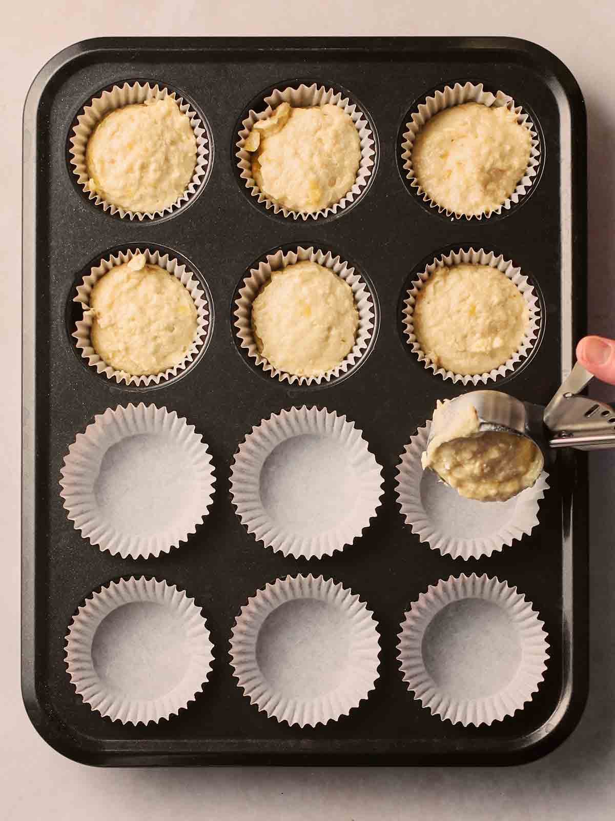 A muffin tray on a white counter, with half of them filled with cupcake mixture, and the other half being filled with batter.