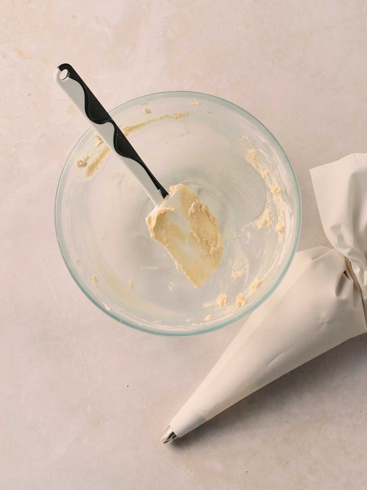 An empty bowl with the remnants of buttercream icing inside with a spatula, with a filled piping bag to the side, on a white surface.