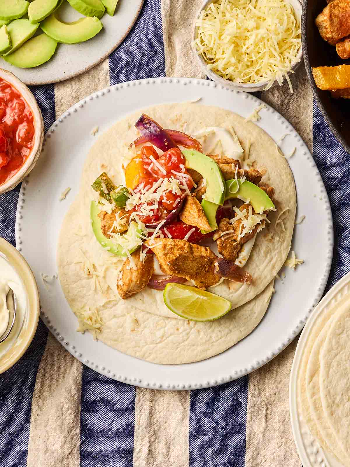 A plate of chicken fajitas in a wrap with lots of toppings, with lots of side dishes surrounding the plate, ready to eat.