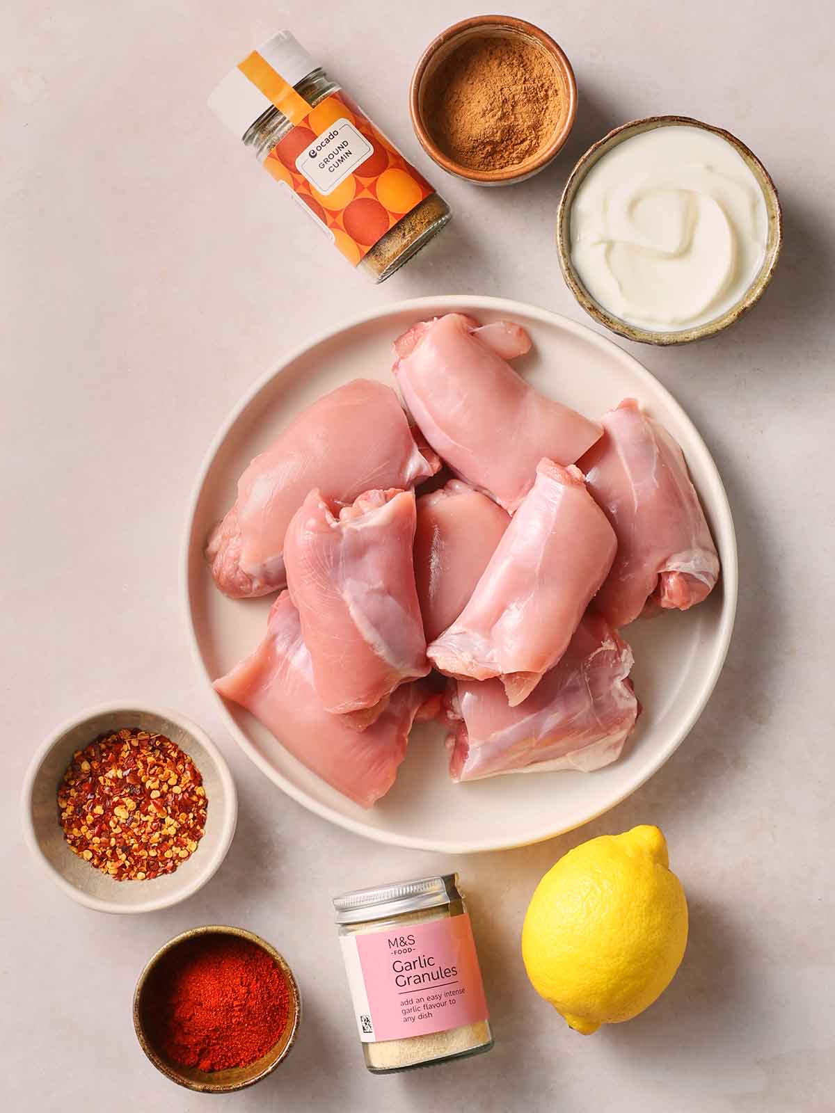 The raw ingredients for making Chicken Kebabs laid out on a white counter, including chicken thighs, spices and natural yoghurt.