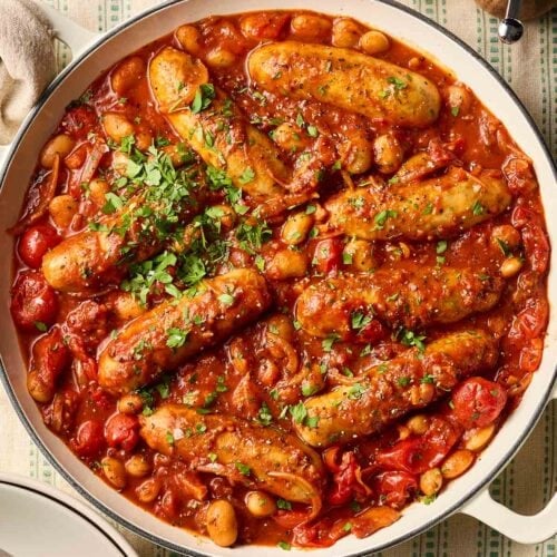 A big pan of sausage and bean casserole with tomato sauce on a dining table with a stripy green tablecloth, ready to serve.