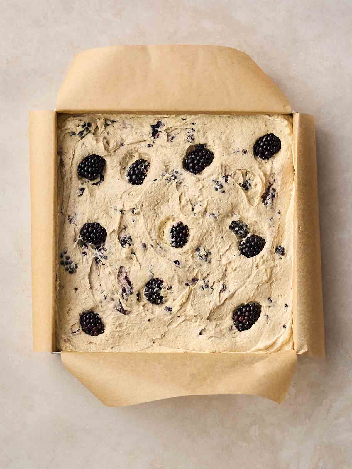 A lined square cake tin on a white surface, with unbaked Blackberry Cake mixture inside.