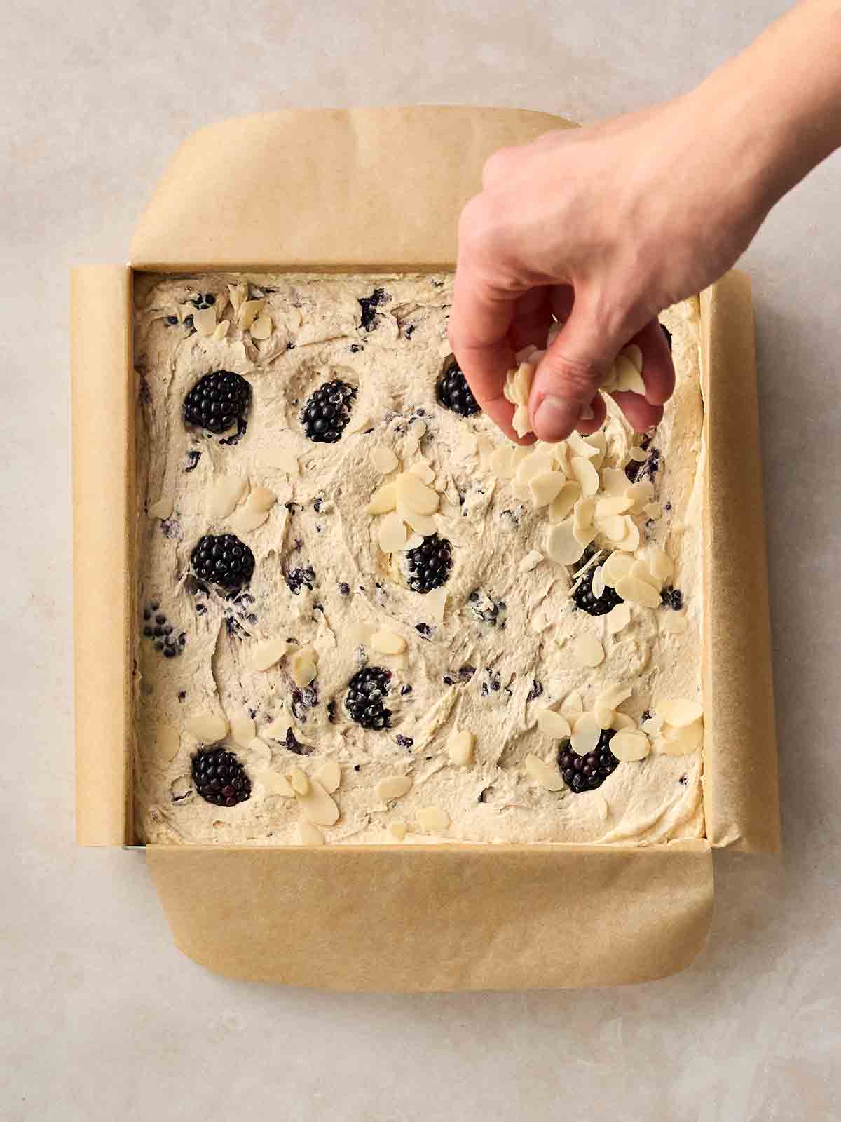 A hadn't sprinkling flaked almonds over an unbaked Blackberry Cake in a lined, square tin on a white surface.